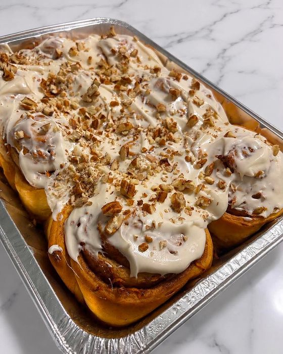 A tray of freshly baked cinnamon rolls topped with swirled cream cheese frosting and sprinkled with chopped pecans.